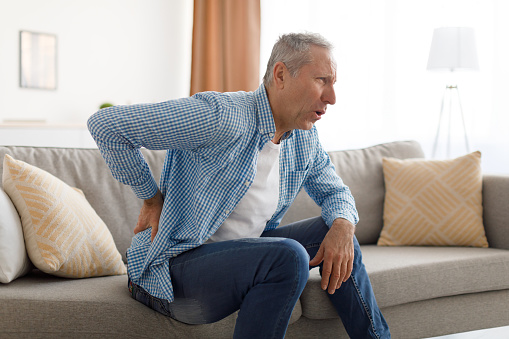 People, Healthcare and Treatment. Portrait of sick mature man suffering from acute side back pain, sitting on couch at home, person touching lower back aching area with hand. Health Problem Concept