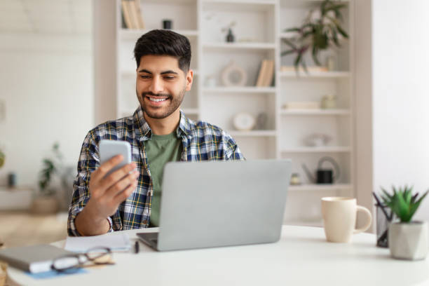 istock Portrait of smiling man using smartphone and pc at home 1476722003