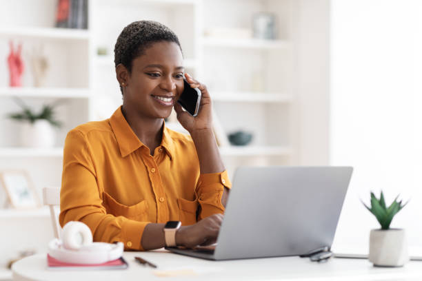 istock Black woman sitting at table with laptop, having phone conversation 1476722082