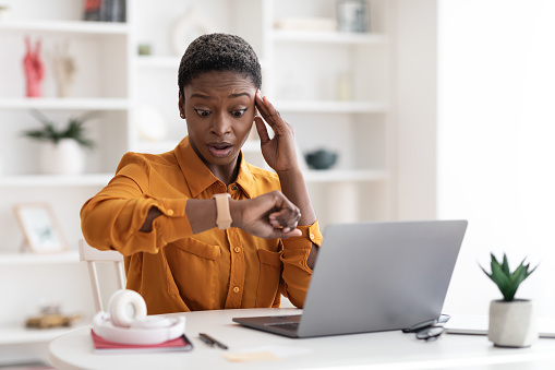 Attractive young african american business woman sitting at workdesk in front of laptop, working online, looking at watch and touching head, got late for business meeting, office interior, copy space