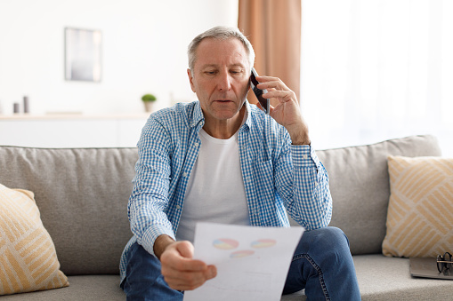 Mature Male Entrepreneur Talking On Phone Holding Paper And Discussing Document Working From Home Sitting On The Couch. Mobile Communication, Senior People And Gadgets Concept