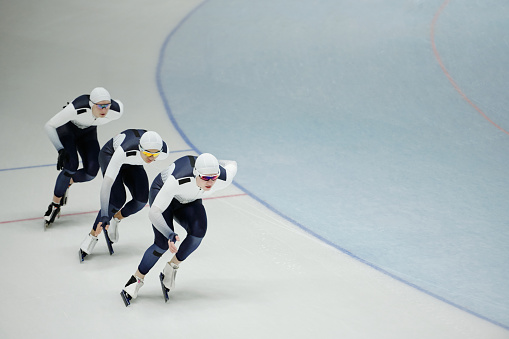 Row of three athletes in sports uniform, skates and eyeglasses sliding forwards down ice rink while taking part in competition or race
