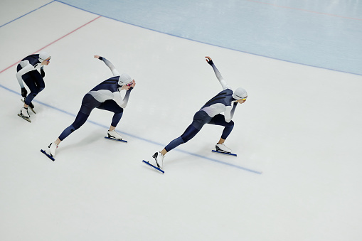 Row of young men in sports uniform and skates training some speed skating exercises while moving forwards along ice rink on arena