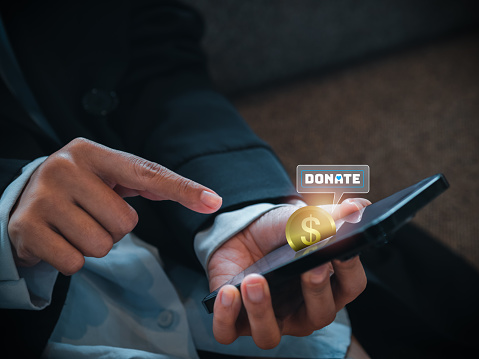 Close-up digital golden coin and donate icon appear on smart mobile phone on donation application or website, pressing by businessperson's hands. Donation online by cellphone app concept.