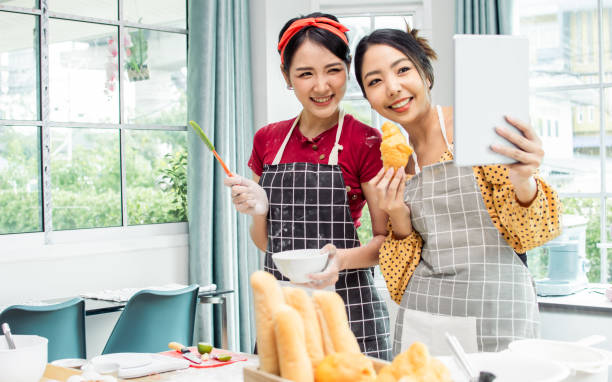 istock Two beautiful Asian women cooking food, making bread for breakfast and bakery while using tablet, streaming live video clip or taking selfie photo together with happiness and fun in kitchen at home. 1476740507