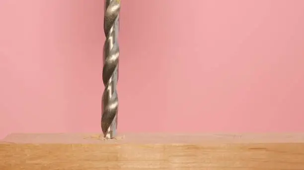 On a pink background, a drill while drilling wood, close-up. A shiny metal drill bit at work. Slow motion