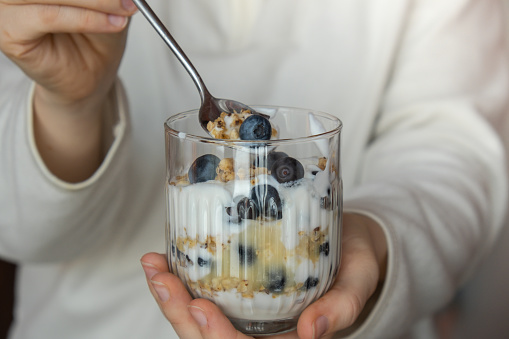 Unrecognizable woman eating Morning granola breakfast with homemade greek yogurt, blueberries in glasses on blue wooden background. Healthy diet Crunchy granola with yogurt nuts and blueberries for healthy light breakfast