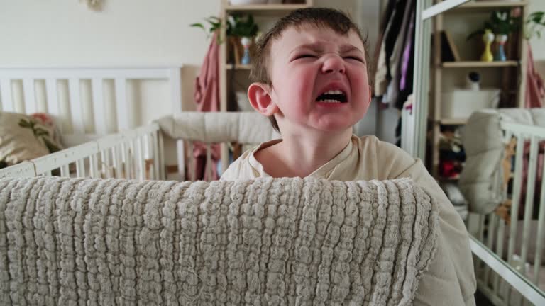 Toddler baby is naughty while standing in the crib. Sleepy child cries without tears, face close-up. Kid age one year nine months