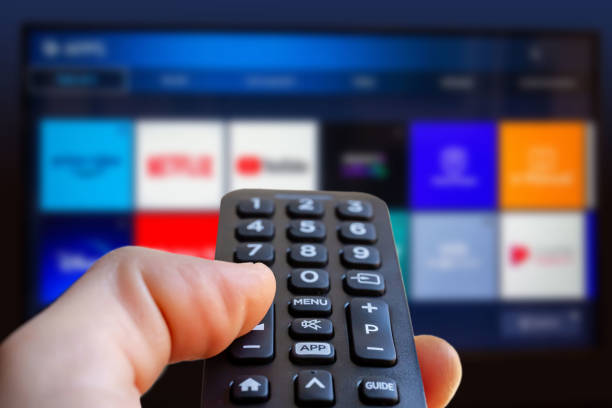 a man is holding a remote control of a smart tv in his hand. in the background you can see the television screen with streaming entertainment apps for video on demand - changing channels imagens e fotografias de stock