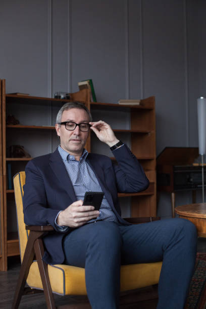 istock Handsome elderly grey haired businessman in glasses looking on his mobile phone, sitting on chair in office 1476754411