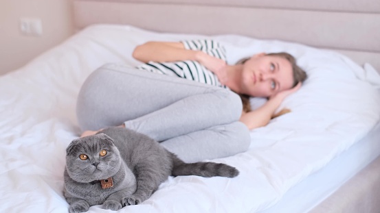 Depressed young woman lying in bed with a beautiful cat sitting next to her, unhappy woman suffering from insomnia or depression, psychological problem, blurred background