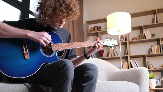 A curly-haired guy is sitting on a sofa and playing an acoustic guitar, he has a headache. Migraine and headache in a young man