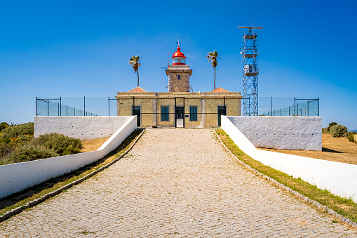Fenced Farol da Ponta da Piedade lighthouse in Lagos captured from the driveway on a sunny day with blue sky, showcasing its tower and gate, creating an almost symmetrical composition with copy space.