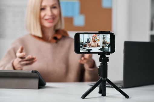 Close-up of smartphone standing on tripod with teacher recording online lesson for her students at table in class