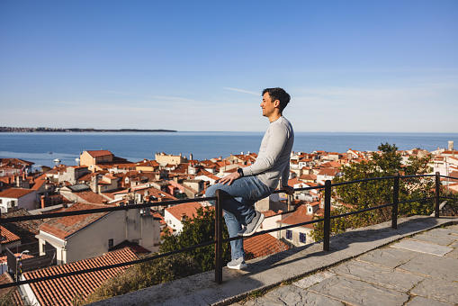 Happy Male Asian Tourist Traveler Sitting On A Fence While Enjoying The View Of The Sea