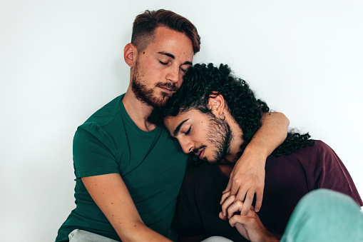 two men hugging each other with closed eyes - same-sex love and affection - hands in hands against white wall - casual man with Mediterranean or North African or Middle Eastern skin