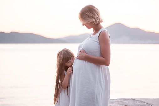 Close-up portrait of little girl hugging pregnant mother. Daughter in white sundress kisses belly to middle aged woman by the sea at sunset. Family travel. The concept of happy motherhood. Copy space