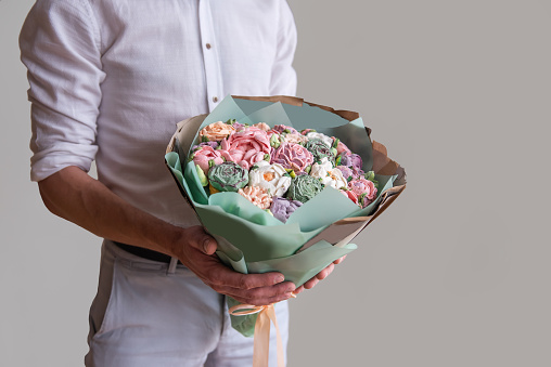 Faceless man holds bouquet of marshmallow sweet flowers in hands. Gift in pastel green wrapper with delicate sugarless roses on gray background. Small business home. Hobby baking. Holiday present