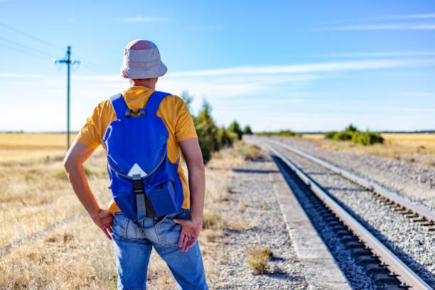 노란색 티셔츠, 파란색 배낭, 모자를 쓴 뒤에서 햇볕이 잘 드는 시골 환경의 기차 선로에서 먼 곳을 바라보고 있는 남자. 기차 트랙을 따라 하이킹 - picardy 뉴스 사진 이미지