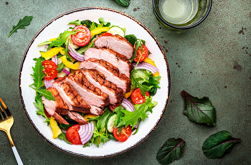 Duck salad with grilled breast and vegetables: red tomato, cucumber, paprika, onoin  and chard, frisse, mizuna and arugula leaves on green table background, top view