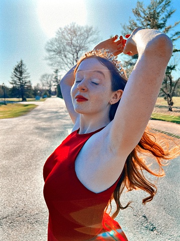 A high school red headed girl on prom night in prom attire outside, closing her eyes and enjoying a moment in the wind and sunshine, her red hair streaming out and wearing a red dress and small gold crown. Pembine, Wisconsin.