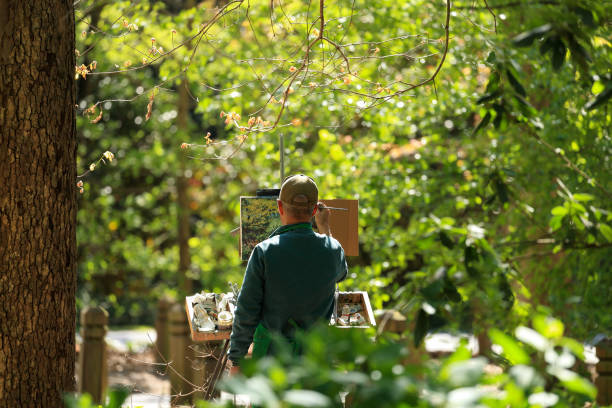 Norfolk, Virginia, USA - April 16, 2021: Artist painting a scene at the local botanical gardens. I like this photograph because the artist is painting a woodland scene and he is dressed in green, almost blending into the scene.