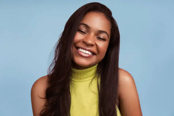 Photo of Delighted young ethnic lady smiling with closed eyes in blue studio