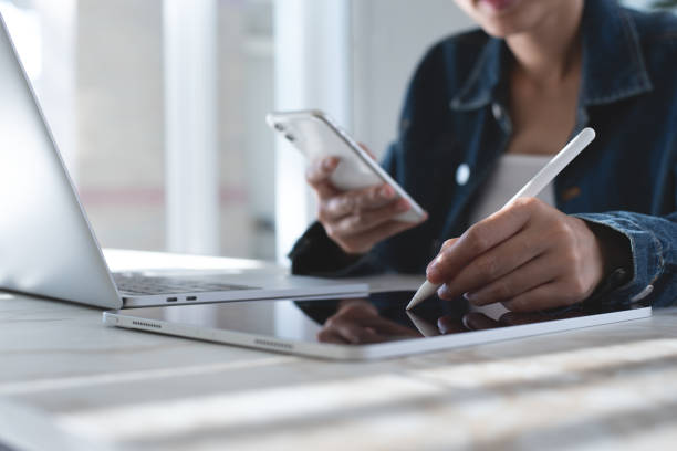 primer plano de una mujer de negocios que usa un lápiz óptico que firma el documento electrónico en una tableta digital, la mano sosteniendo el teléfono móvil mientras trabaja en una computadora portátil en la oficina, la firma electrónica, la firma - working at home calendar laptop women fotografías e imágenes de stock