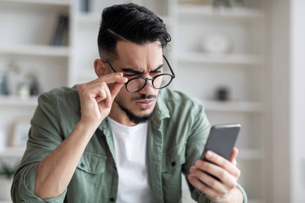 Eyesight Problems Concept. Young Arab Man In Eyeglasses Looking At Smartphone Screen Eyesight Problems Concept. Young Arab Man In Eyeglasses Looking At Smartphone Screen And Frowning, Millennial Guy Trying To Read Message, Suffering From Astigmatism And Bad Vision, Closeup Shot squint eyes stock pictures, royalty-free photos & images