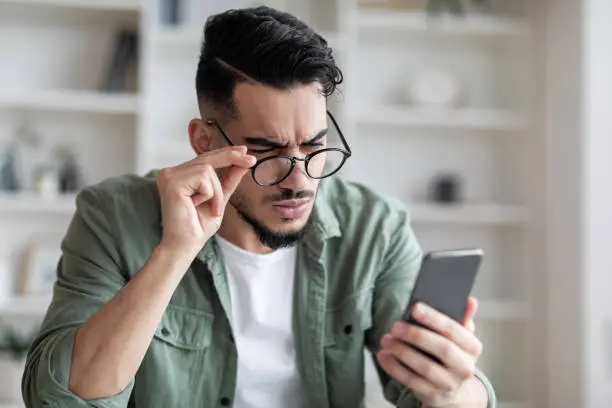 Photo of Eyesight Problems Concept. Young Arab Man In Eyeglasses Looking At Smartphone Screen