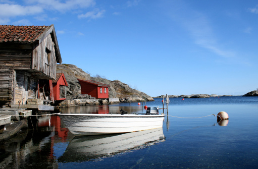A blue sky, sunny day with bright, beautiful colours, as old wooden cabins nestle along the Norwegian fishing harbour. A small boat rests in the foreground on a calm sea - No colour enhancement, it really is that blue in the North!