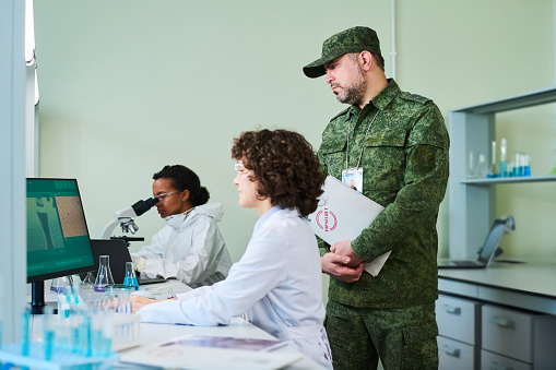 Mature man in camouflage uniform standing by female clinician while both looking at computer screen during secret research in laboratory