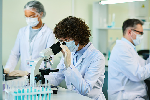 Mature brunette woman in lab coat and mask looking in microscope while studying cell of new virus by workplace among colleagues