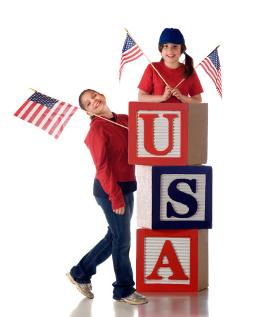 Two girls in red and blue displaying flags while leaning on oversized USA alphabet blocks.  Isolated on white.