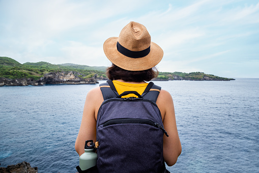 Back view of woman traveler looking at the ocean on vacation trip to Nusa Penida, Bali. Female with backpack enjoying freedom in nature. Traveling, healthy lifestyle and wellness concept.