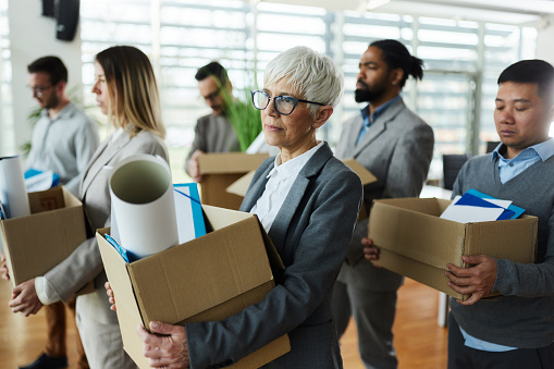 Large group of displeased business people carrying carton boxed with their belongings after being fired from their jobs. Focus is on senior woman.