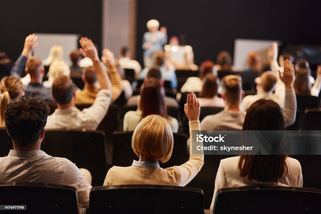 Back view of crowd of people raising hands on a seminar in convention center. Rear view of large group of people raising their arms to ask questions on press conference in convention center. Meeting Stock Photo