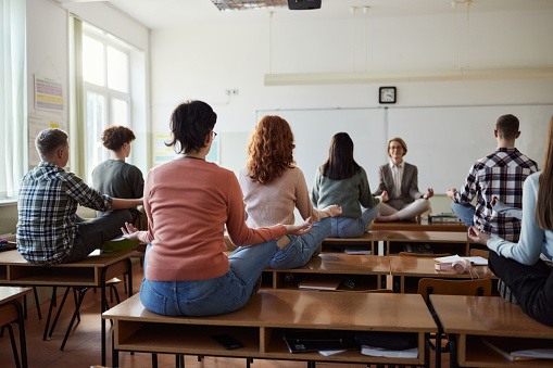 Rear view of large group of high school students and their teacher meditating in Lotus position during a class in the classroom.
