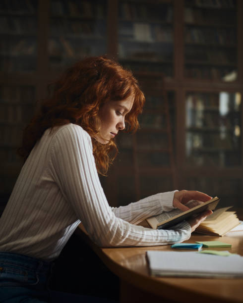 istock Redhead female student reading a book in library. 1476981919