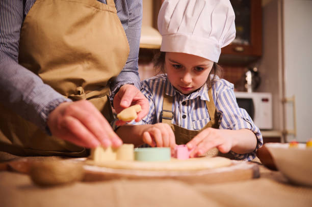 istock Little girl in chef hat prepares Easter gingerbread biscuits, cuts dough using cookie cutters, under her mom's guidance 1476983865