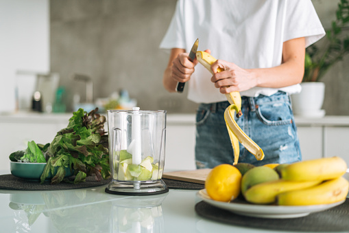 Young slim woman in white t-shirt and blue jeans cooking smoothie with bananas healthy food in kitchen at home