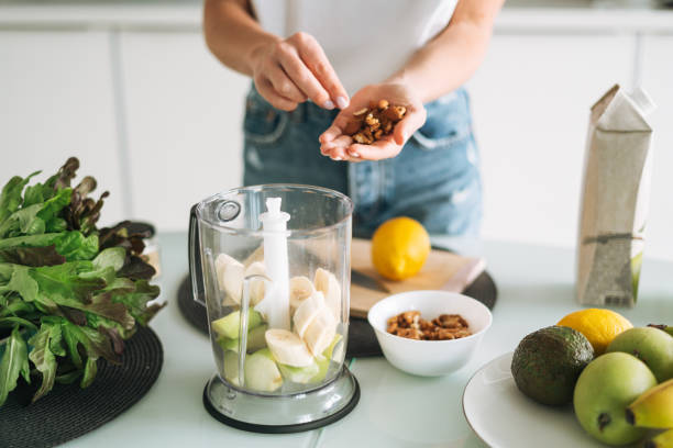 junge schlanke frau in weißem t-shirt und blauer jeans kocht smoothie mit bananen gesundes essen in der küche zu hause - routine foods and drinks clothing household equipment stock-fotos und bilder