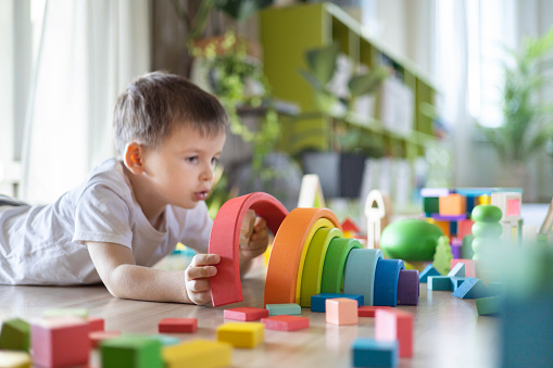 Funny baby boy playing multicolored arch Montessori wooden material structure building closeup. Little kid construction curved colored blocks on floor at childish room early development education