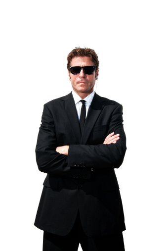 A man in a dark suit, with dark sunglasses, arms crossed and ear piece in.  He is standing guard and serious.  Could be a secret service agent, a security guard or just some tough, stern looking businessman.