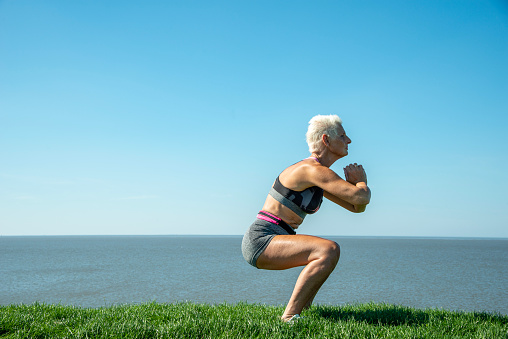 Senior woman exercising outside in the grass on a sunny day. Sea in the background.