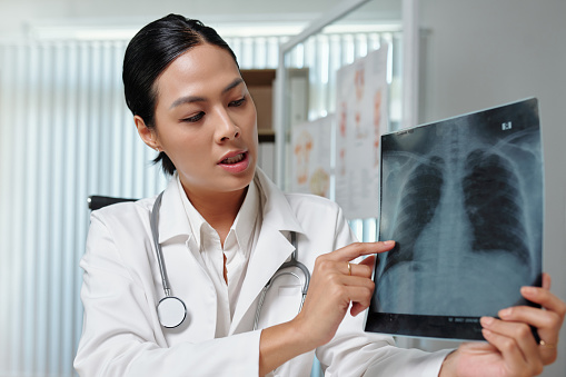 Young female radiologist in lab coat commenting lung x-ray to patient during online consultation while sitting in medical office