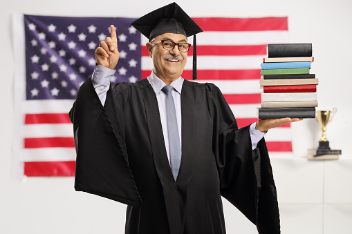 Smiling mature man in a graduation gown holding a pile of books and pointing in front of a USA flag