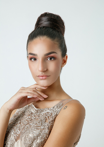 Portrait of beautiful young brunette with hair in bun, wearing champagne colored sleeveless top and looking at camera with hand on chin, studio shot