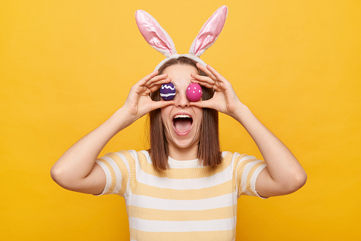 Indoor shot of amazed surprised overjoyed woman wearing rabbit ears covering eyes with Easter eggs, screaming with happiness, celebrating holiday isolated on yellow background