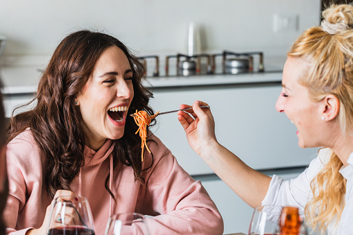 Couple of young girls friends laughing having fun joking with spaghetti. Blonde girl feeding her brunette friend with a fork. Playful moment in an intimate lunch at home